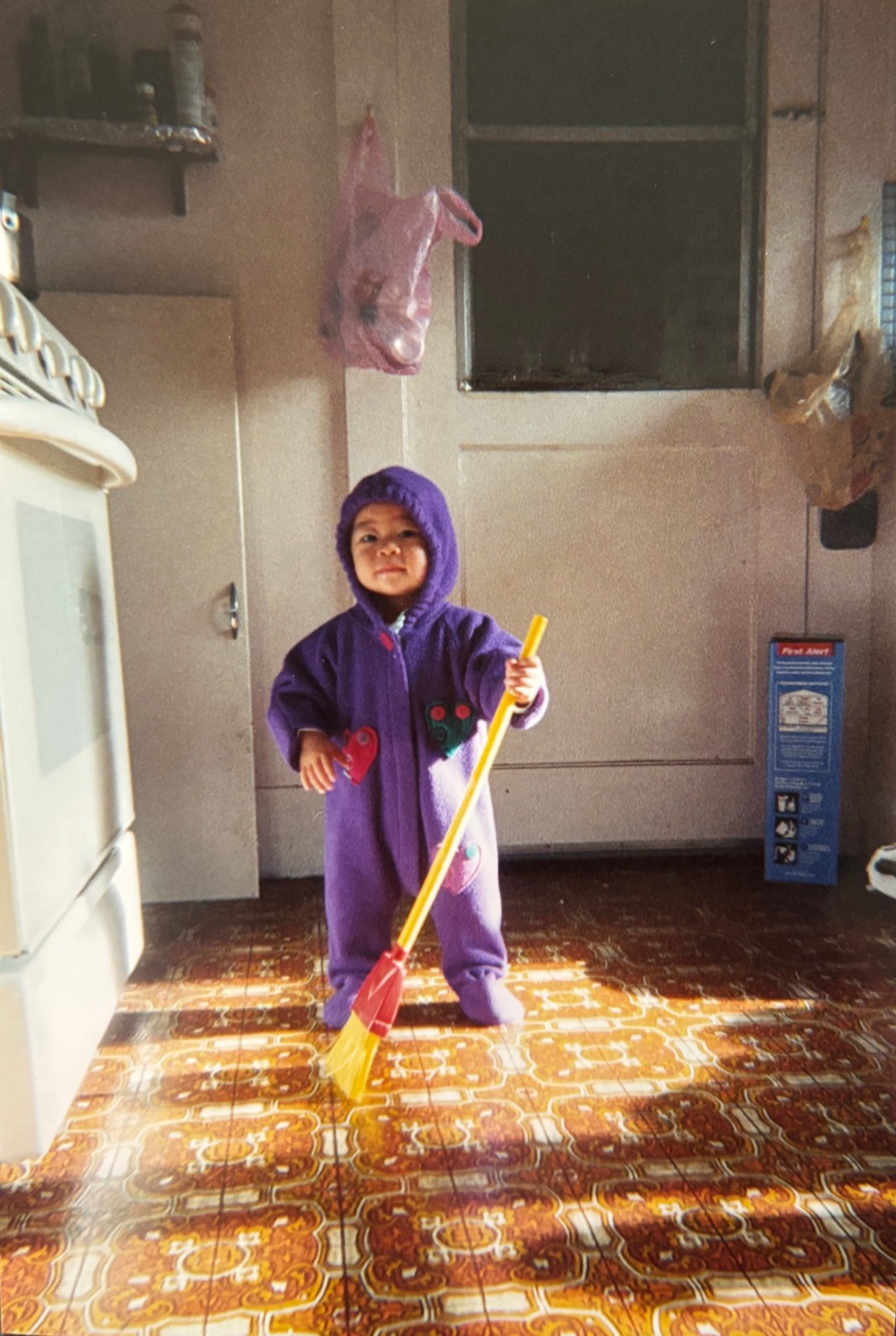 Claudia as a toddler in a purple snowsuit, sweeping the kitchen floor with a toy broom